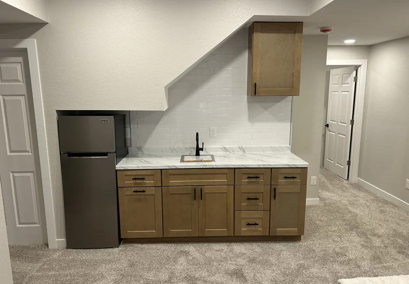 Wood kitchenette cabinets with black pulls, a marble-look counter and a white subway tile backsplash.